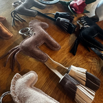 A collection of handmade leather keychains and small accessories arranged on a wooden tabletop. The set includes several horse-shaped keychains in taupe, brown, and blush pink, along with black leather gloves, a dark green rose keychain, and leather tassels.