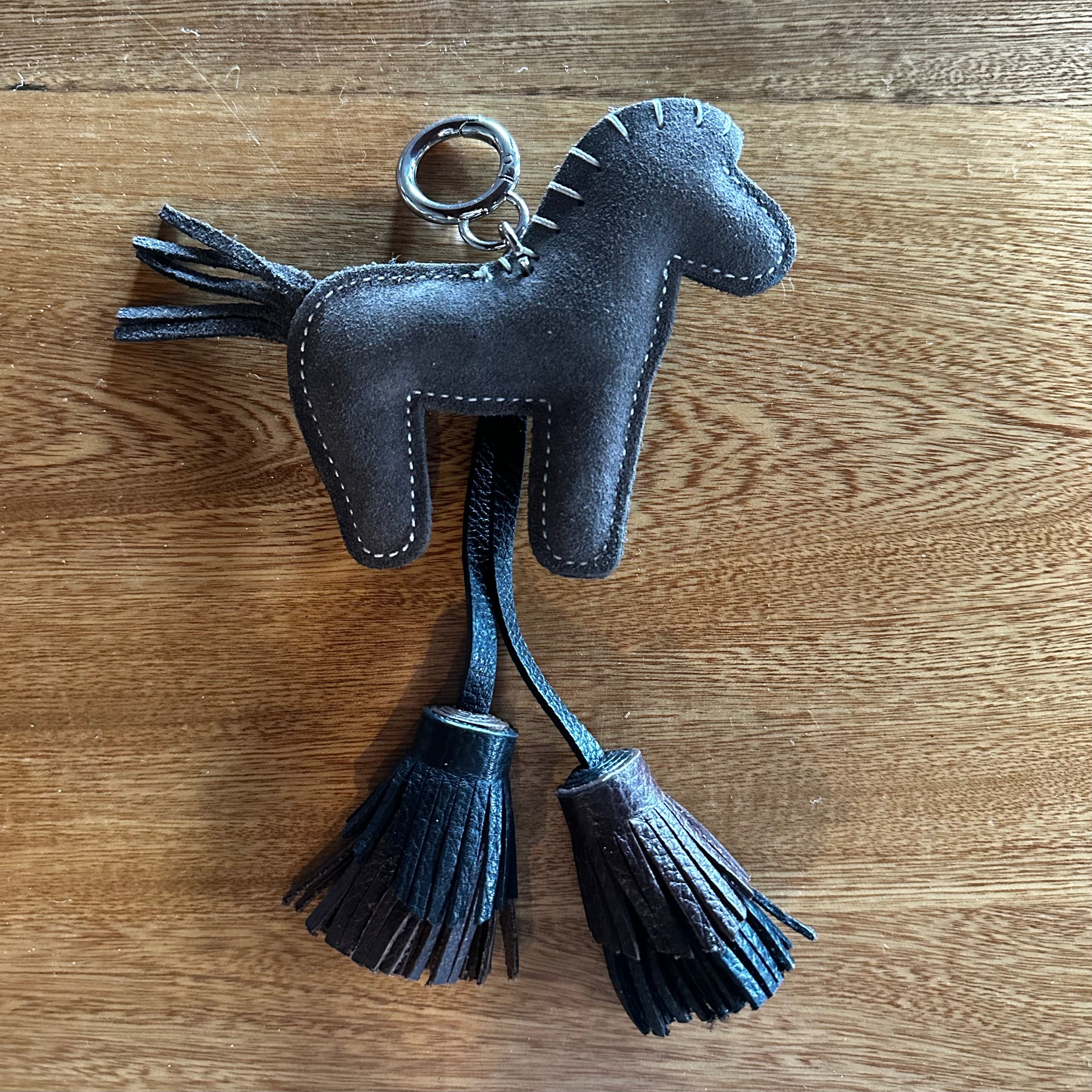 A dark brown leather horse-shaped keychain with two hanging tassels and a metal key ring, placed on a wooden surface.