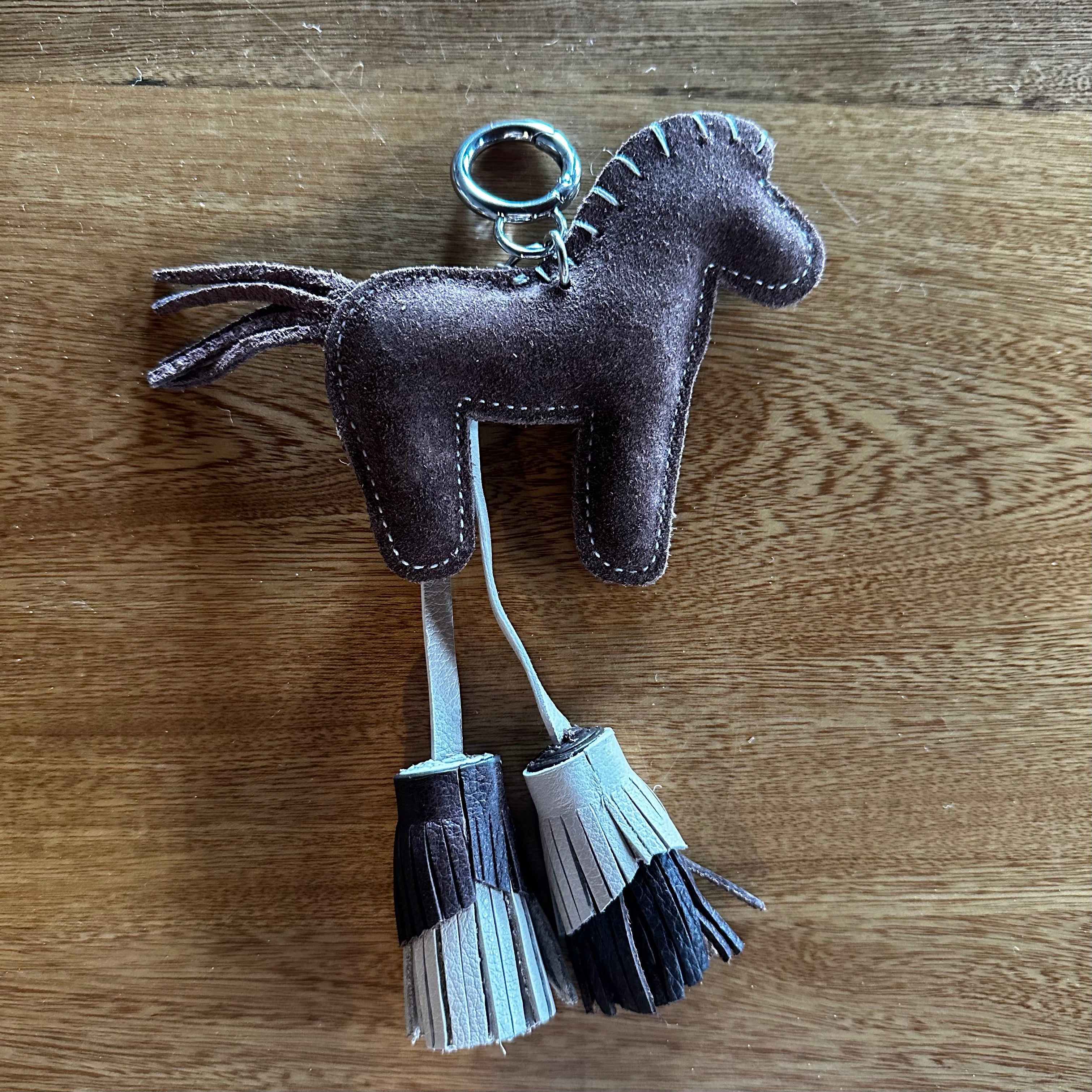 A small horse-shaped keychain or bag charm made from dark brown suede-like material, with a metal key ring and two dangling fringe tassels in dark brown and light beige, placed on a wooden surface.