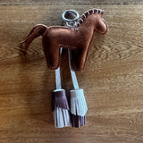 A brown leather horse-shaped keychain with white stitching, a silver metal key ring, and two hanging fringe tassels in white and dark brown, displayed on a wooden surface.