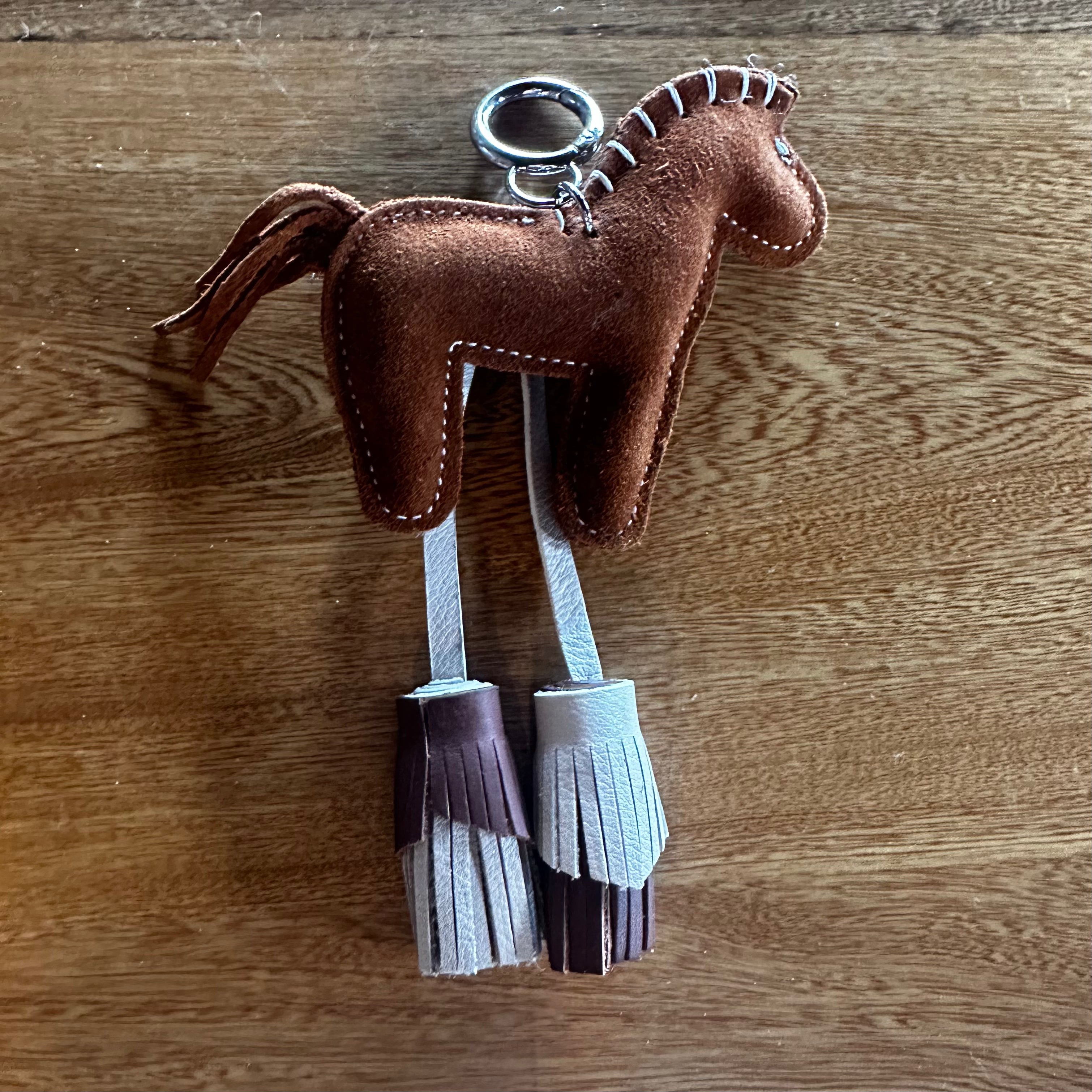 A brown leather horse-shaped keychain with white stitching, a silver metal key ring, and two hanging fringe tassels in white and dark brown, displayed on a wooden surface.
