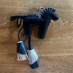 A black felt horse-shaped keychain or bag charm with white stitching, hanging tassels, and a silver metal key ring, placed on a wooden surface.