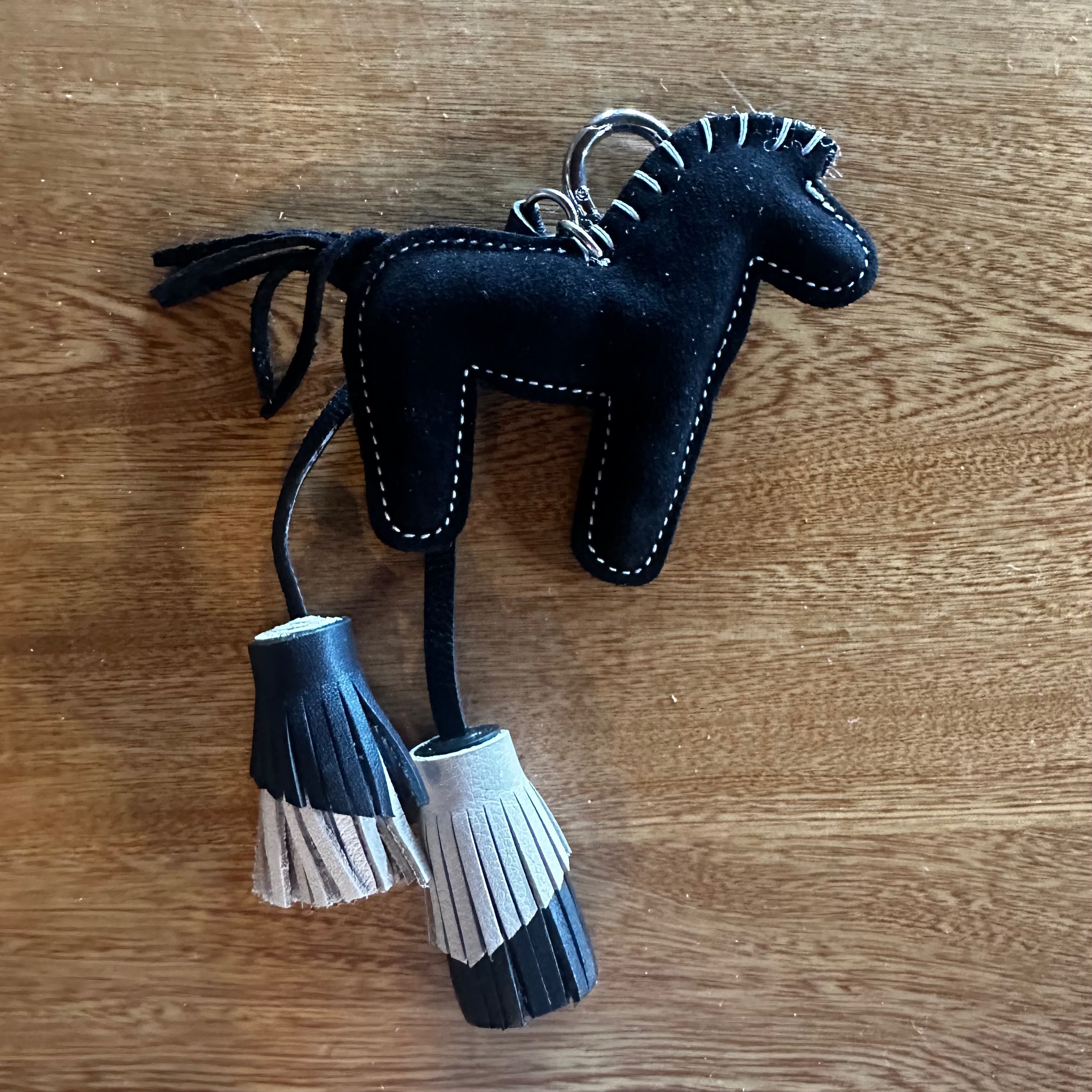 A black felt horse-shaped keychain or bag charm with white stitching, hanging tassels, and a silver metal key ring, placed on a wooden surface.