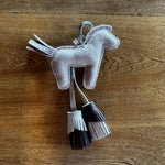 A small suede horse-shaped keychain or bag charm in pale lavender-gray, with two dangling fringe tassels in black, white, and gray, photographed on a wooden surface.