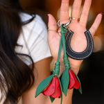 A close-up of a woman's hand holding a leather keychain set featuring a black horseshoe and two red rose-shaped keychains with green stems, against a blurred dark background.