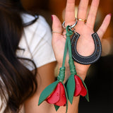A close-up of a woman's hand holding a leather keychain set featuring a black horseshoe and two red rose-shaped keychains with green stems, against a blurred dark background.