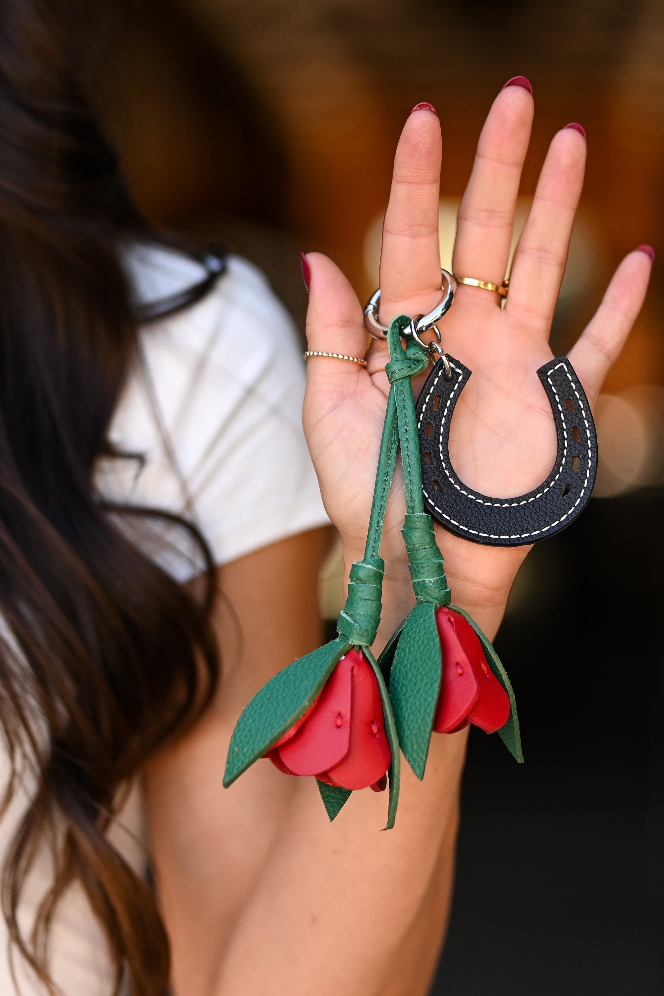 A close-up of a woman's hand holding a leather keychain set featuring a black horseshoe and two red rose-shaped keychains with green stems, against a blurred dark background.