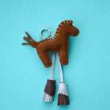 A brown suede horse-shaped keychain with white and brown fringe tassels, attached to a silver metal key ring, set against a solid light blue background.