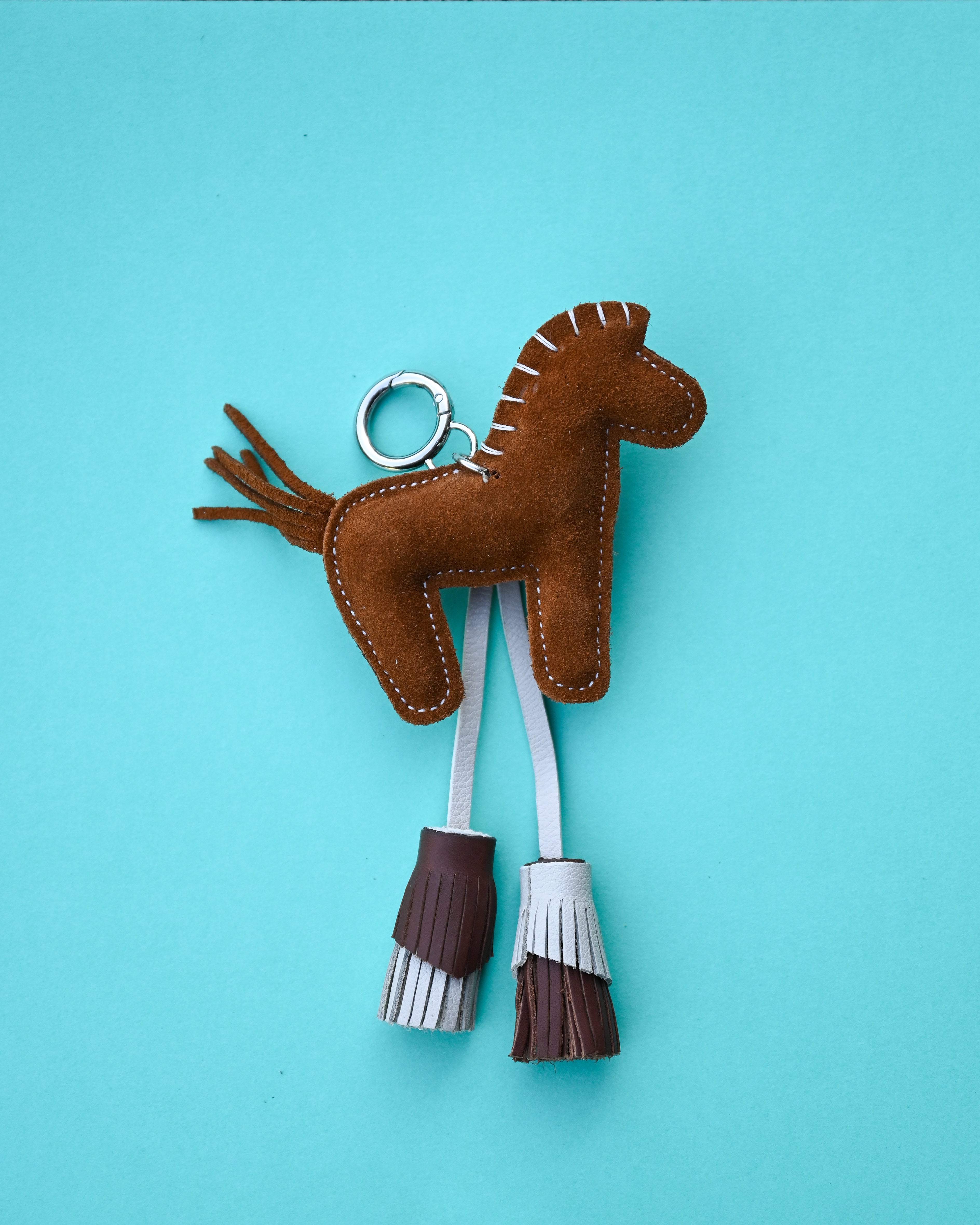 A brown suede horse-shaped keychain with white and brown fringe tassels, attached to a silver metal key ring, set against a solid light blue background.