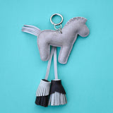 A light gray felt horse-shaped keychain with a silver metal key ring and two hanging tassels, one gray and one black-and-gray, set against a solid turquoise background.
