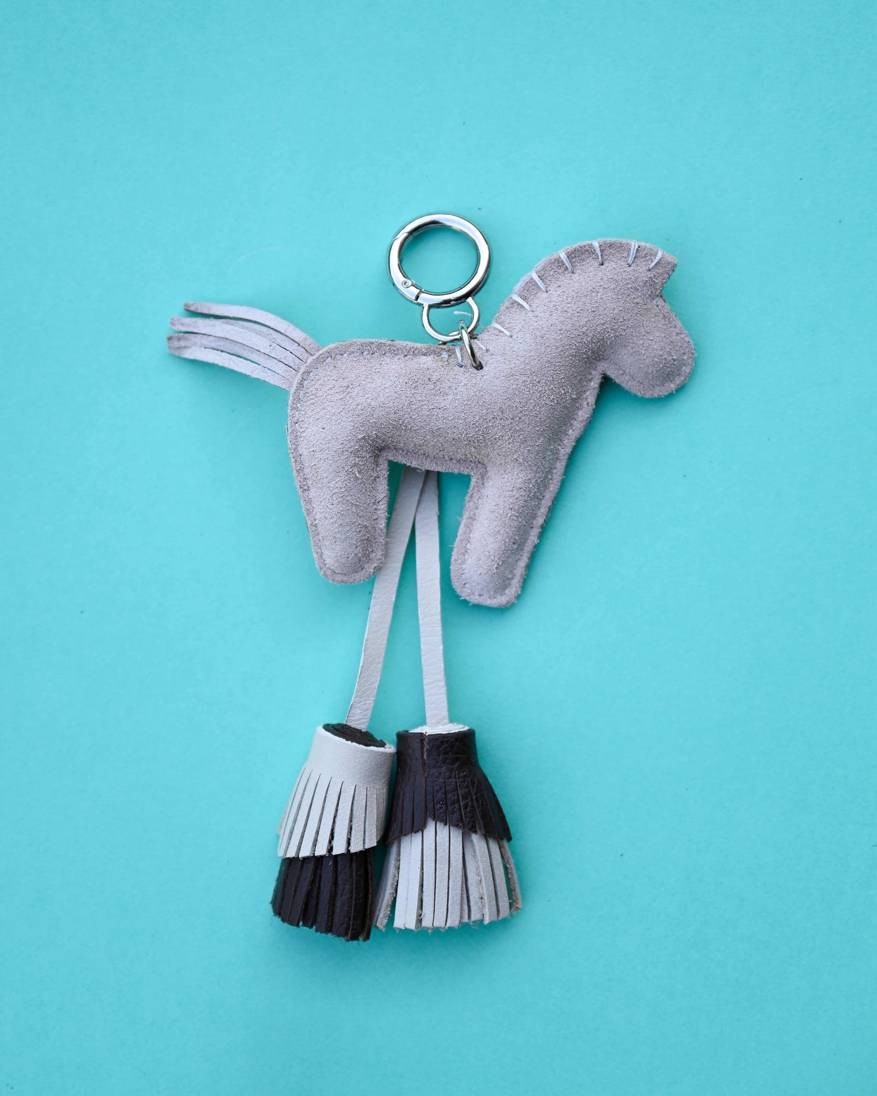 A light gray felt horse-shaped keychain with a silver metal key ring and two hanging tassels, one gray and one black-and-gray, set against a solid turquoise background.