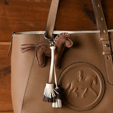 A brown pebbled leather tote bag with a matching leather horse-shaped charm and tassel keychain, displayed on a dark wood background.