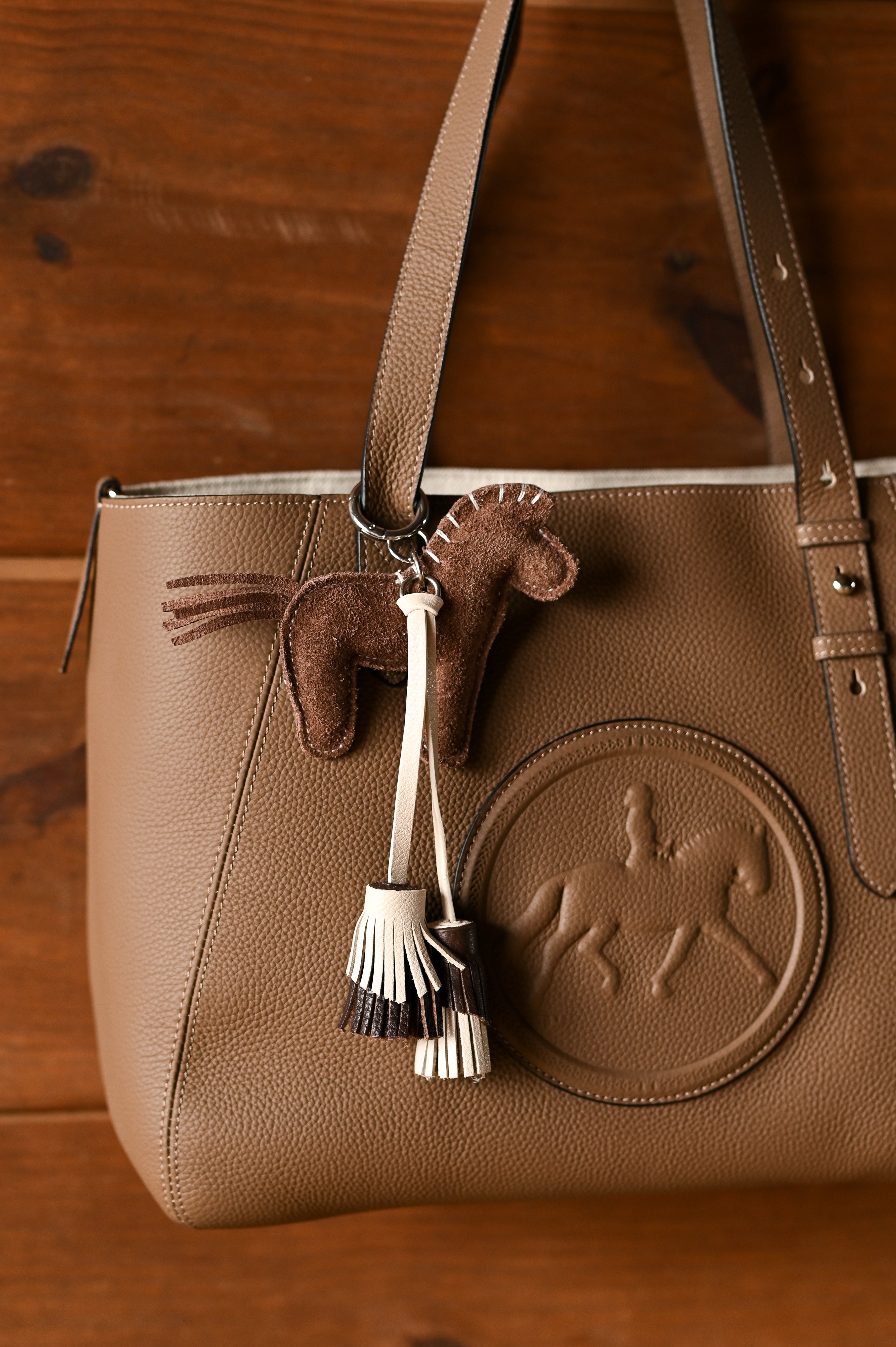 A brown pebbled leather tote bag with a matching leather horse-shaped charm and tassel keychain, displayed on a dark wood background.
