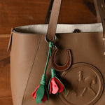 A brown pebbled leather tote bag with a matching leather handle, featuring a hanging charm with a brown leather horseshoe and red and green flower tassels, set against a dark wood background.